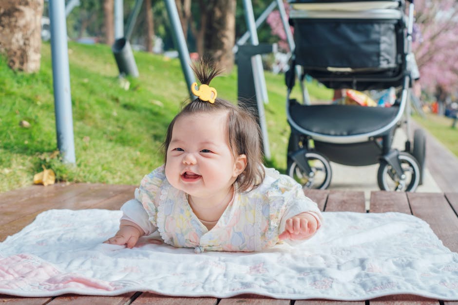 Smiling Asian baby lying on a blanket outdoors beside a stroller on a sunny day.