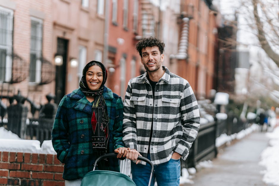 Cheerful ethnic couple in casual wear standing with stroller on sidewalk in snowy town