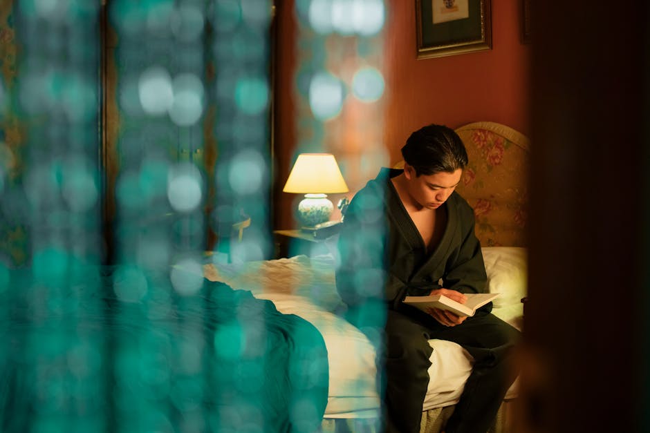 Young man in a robe reading a book in a warmly lit hotel room, blurred beads in the foreground.