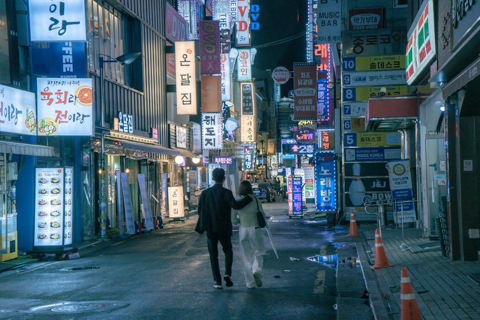 An Asian couple walks through the vibrant, neon-lit streets of Seoul at night, surrounded by shops and signs.