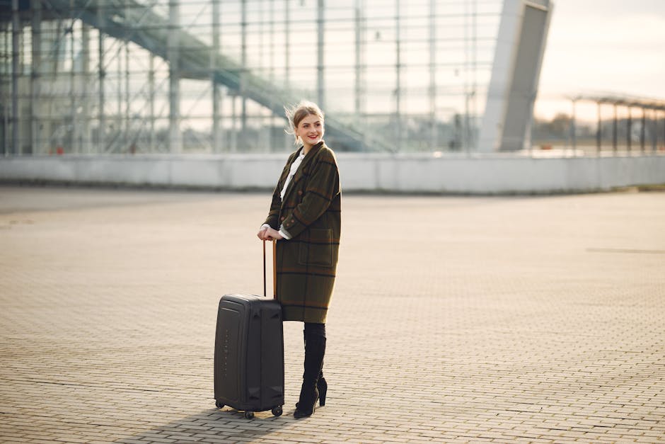 Full length of young joyful woman in outwear and black high heeled boots with luggage standing against glass wall of airport building and looking away