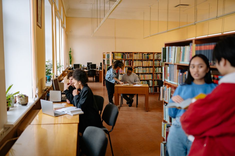 Students studying and collaborating in a university library setting with bookshelves and desks.