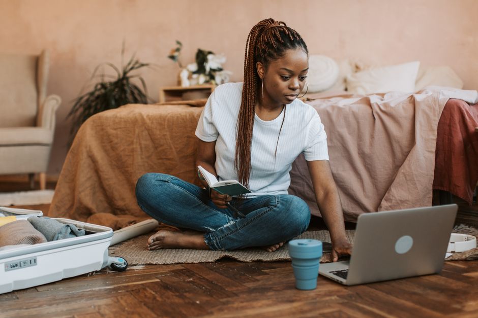 Woman sits on floor packing suitcase, using laptop, and holding notebook in a cozy bedroom environment.