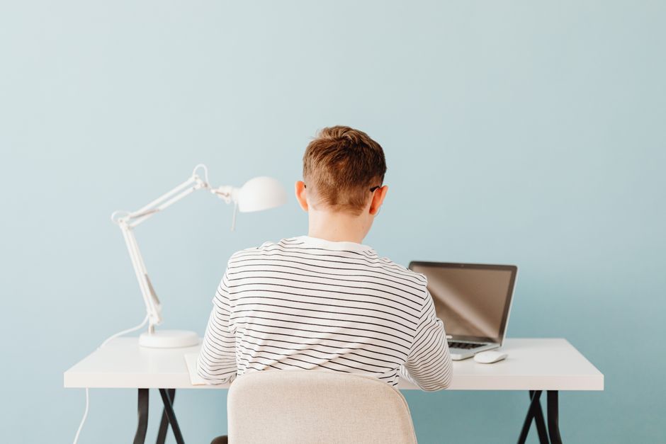 Teenager using laptop at desk under lamp in a minimalist setting with blue background.