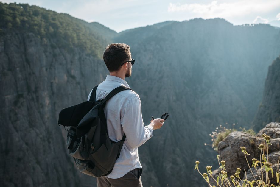 A man with a backpack and compass stands on a cliff, gazing at a scenic gorge.