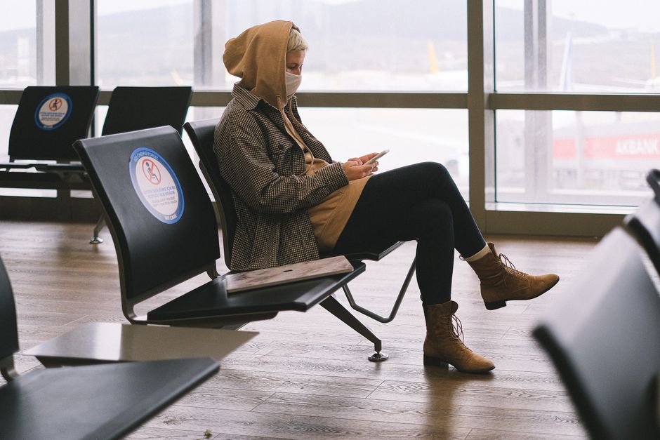 A woman in a hoodie sits in an airport lounge, practicing social distancing while using her phone.